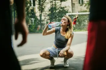 Körperliche Fitness ist Selbstfürsorge: Frau in Sportbekleidung trinkt hockend aus einer Wasserflasche.
