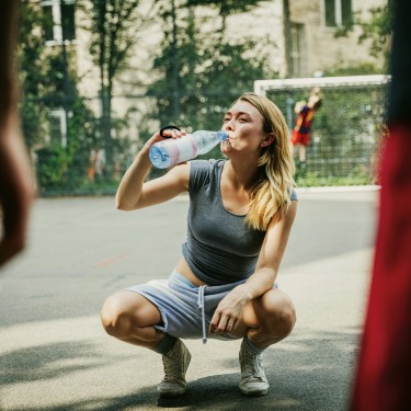 Körperliche Fitness ist Selbstfürsorge: Frau in Sportbekleidung trinkt hockend aus einer Wasserflasche.
