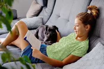 Junge Frau liegt mit einem Buch und einem Hund auf einem Sofa