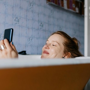 Junge Frau liegt in der Badewanne und hält ein Handy in der Hand