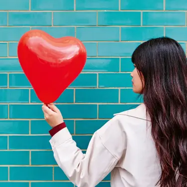 Frau schaut auf Herzform Ballon vor türkisfarbenen Backsteinwand